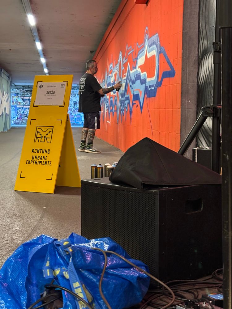 graffitisprayer bei der arbeit in einer unterführung
davor spraydosen subwoofer blaue tasche mit kabeln und ein gitarrenpedalboard