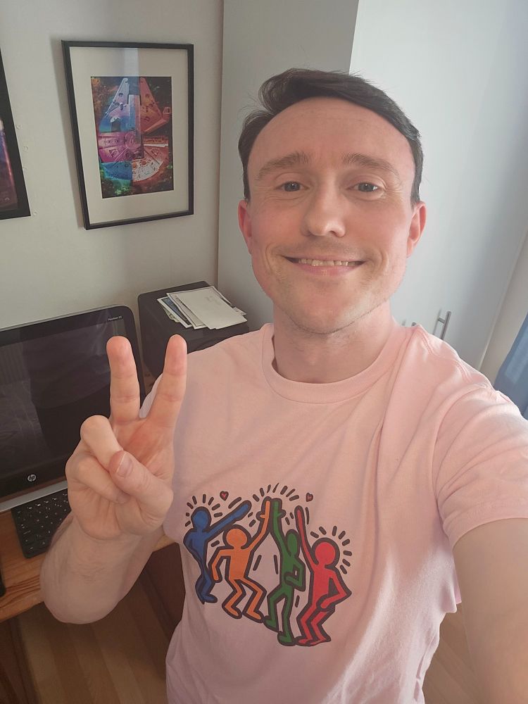 David is in his bedroom wearing a Keith Haring inspired graphic t-shirt smiling at the camera making a peace sign