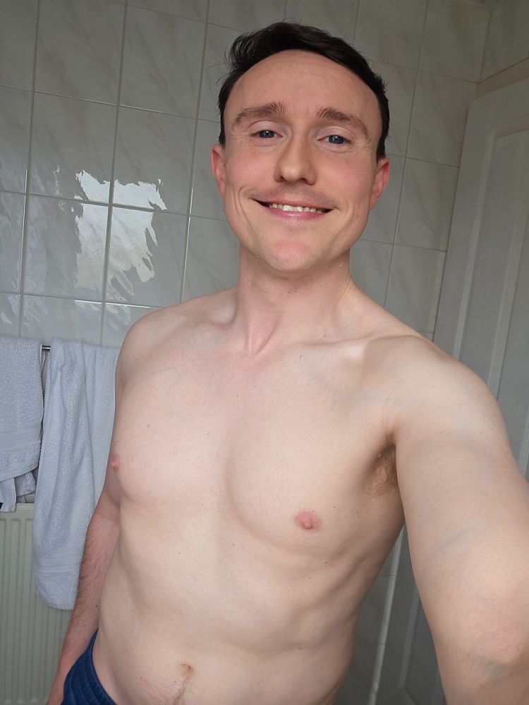 David is standing shirtless in his bathroom wearing blue joggers smiling at the camera 