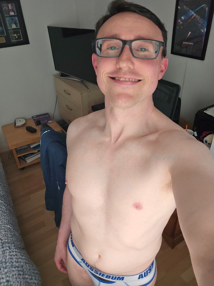 David is standing in his room shirtless pointing the camera slightly upwards and smiling 