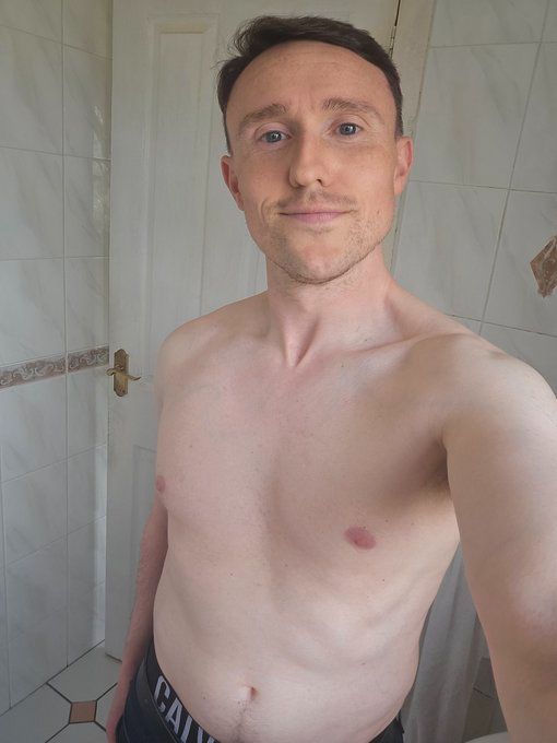 David is standing shirtless in his bathroom in jeans and Calvin Klein underwear holding the camera with a slight smile 
