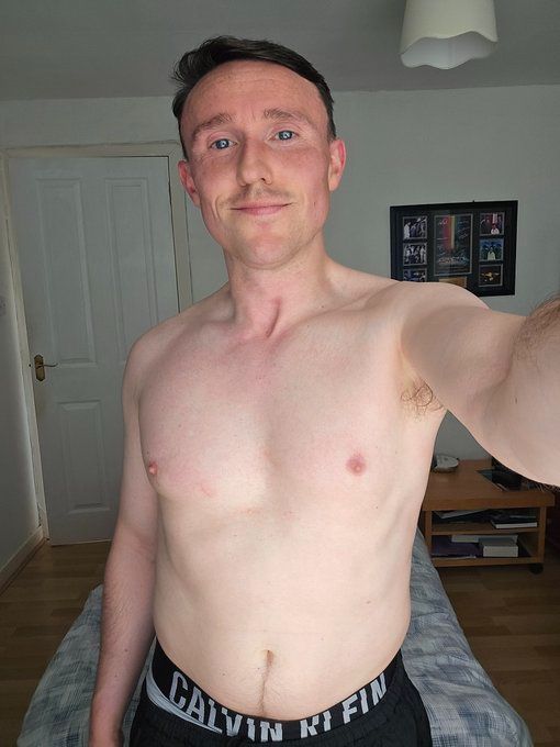 David is standing shirtless in his bedroom wearing shorts and Calvin Klein underwear holding the camera with a slight smile