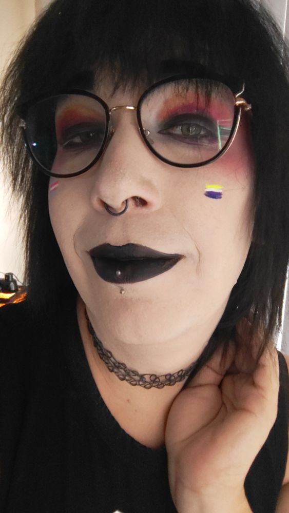 Goth woman with lesbian pride color eye shadow, black lipstick and non binary pride flag painted on her face.