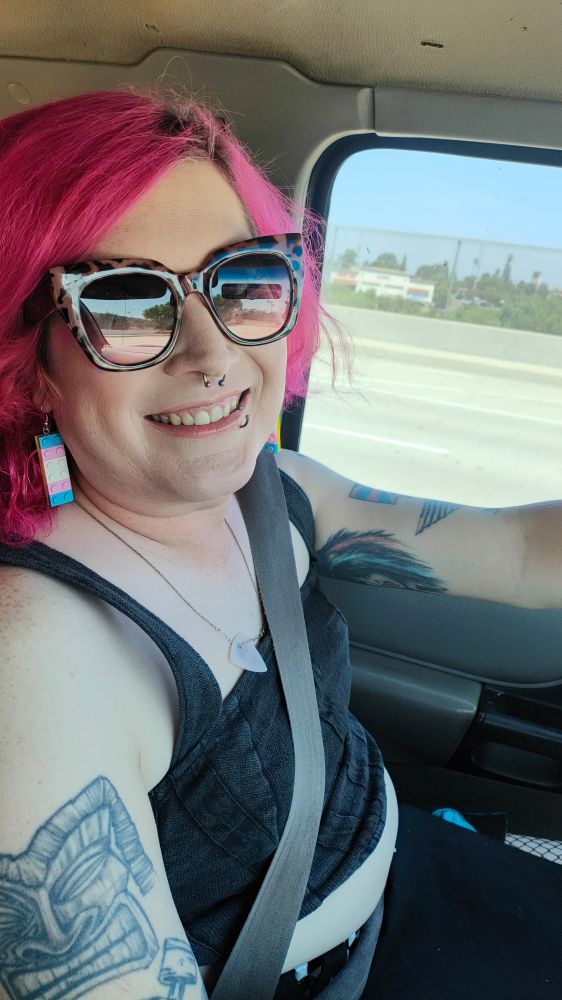 Woman driving with pink hair, sunglasses and smiling.