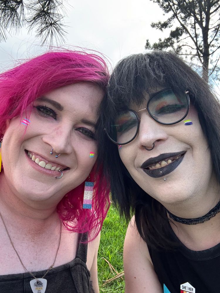 A couple, smiling, one girl with pink hair and the other with black.