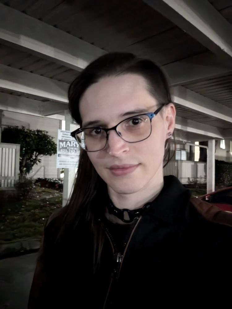Nighttime selfie of Jae from fall 2024. She is standing underneath a parking covering wearing blue framed glasses, with her hair down just past shoulder length, and wearing a black jacket with brown faux leather arms, and a black leather collar with three O-rings.