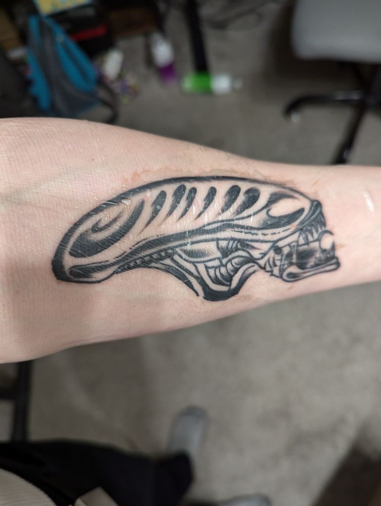 Black and white tattoo of a xenomorph head, from the Alien film franchise, on the inside of Jae's left forearm.