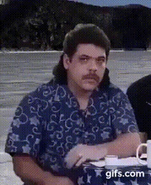 a man with a mullet and mustache is sitting at a table with his hands folded .