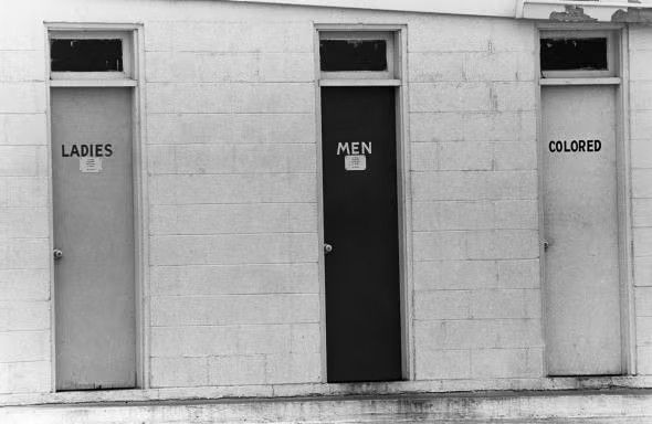 A black and white photograph of a set of three doors to public toilets, designated "Ladies", "Men" and "Colored".