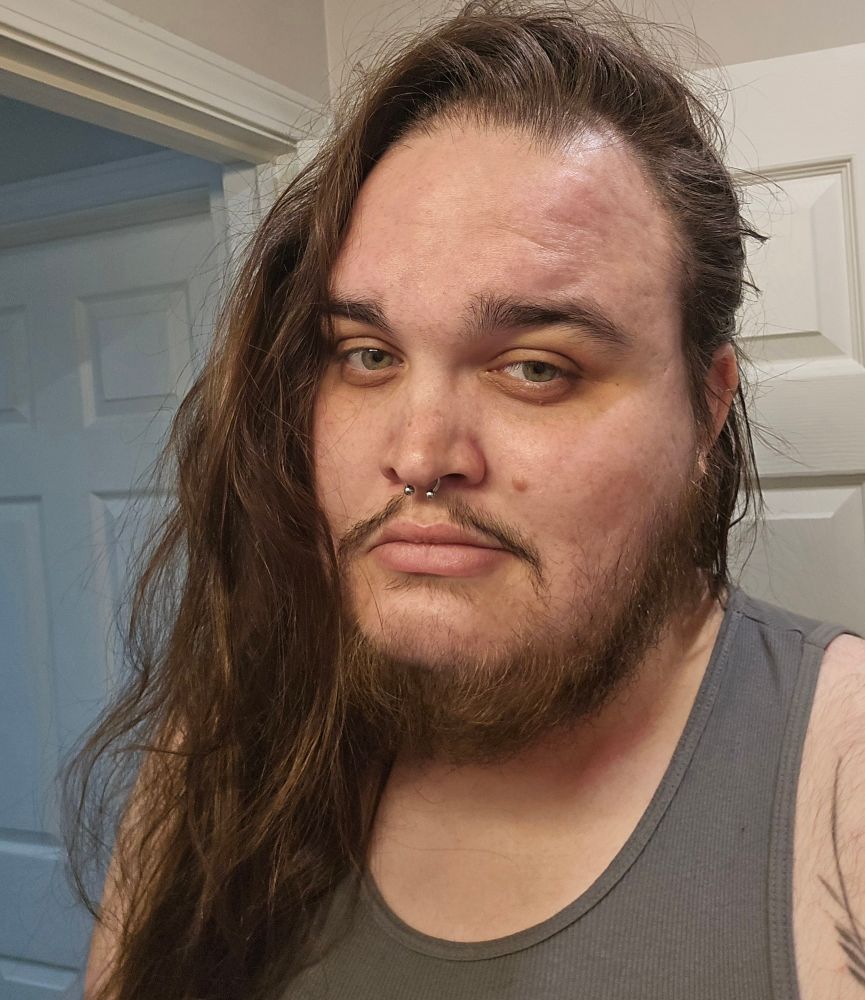 A 28-year-old intersex man (that's me) with long brown hair that hangs down past my chest pushed to one side, green eyes, a septum piercing, and a short beard.