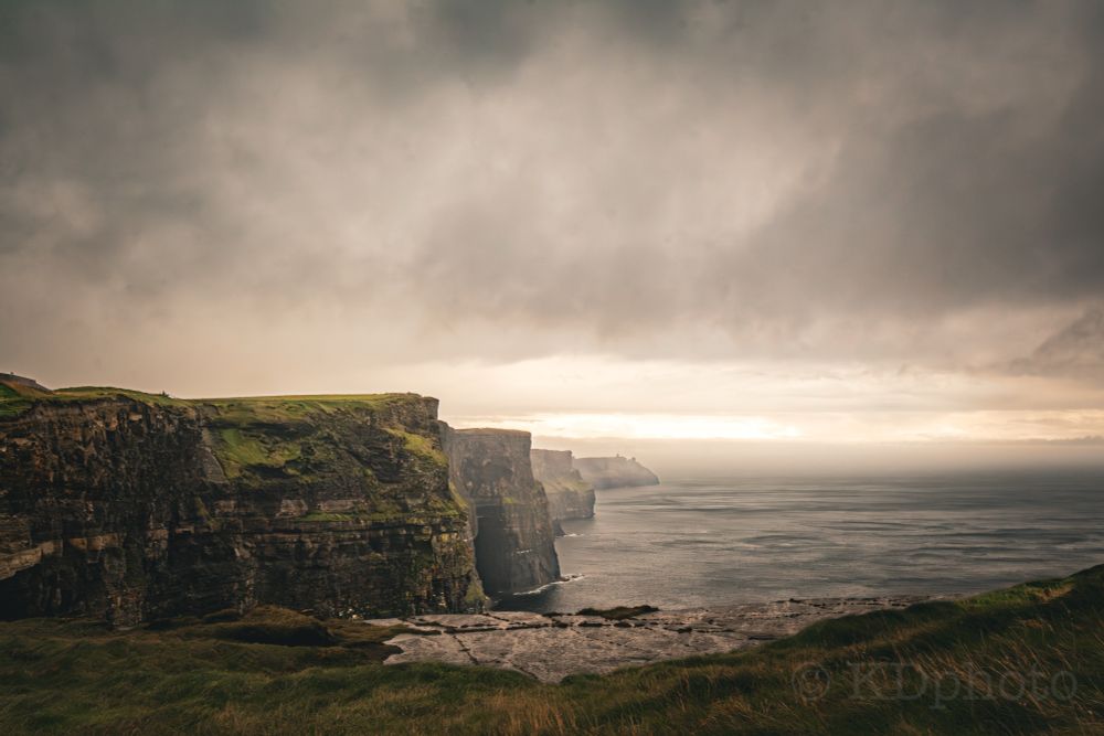 A wide shot landscape photo of the Cliffs of Moher in Ireland. The shot is overlooking from one cliff side and shows a line of cliffs going to the horizon line with dramatic clouds.