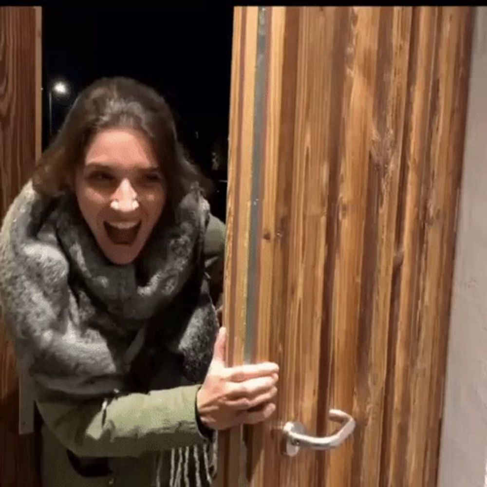 a woman is opening a wooden door with her mouth open and a scarf around her neck .