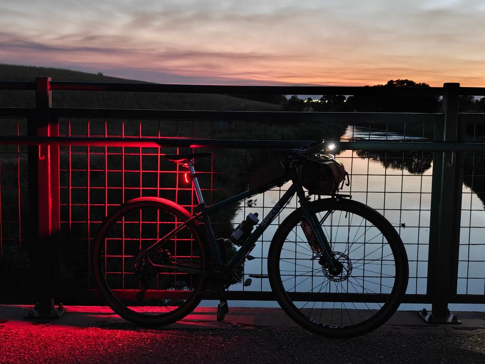 A surly bicycle with a bag attached to the handlebars leans against a metal bridge fence. The fence overlooks a calm river reflecting the pink and orange hues of the sky at sunset.

