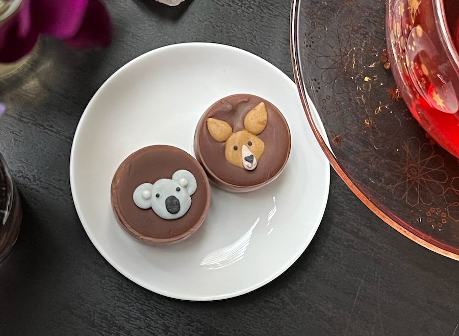 Two small round milk chocolate pralines decorated with sugar faces of a koala (left) and kangaroo (right) on a small round white plate. The edge of a glass saucer sits over the top right edge. Everything sits on a black tabletop. 