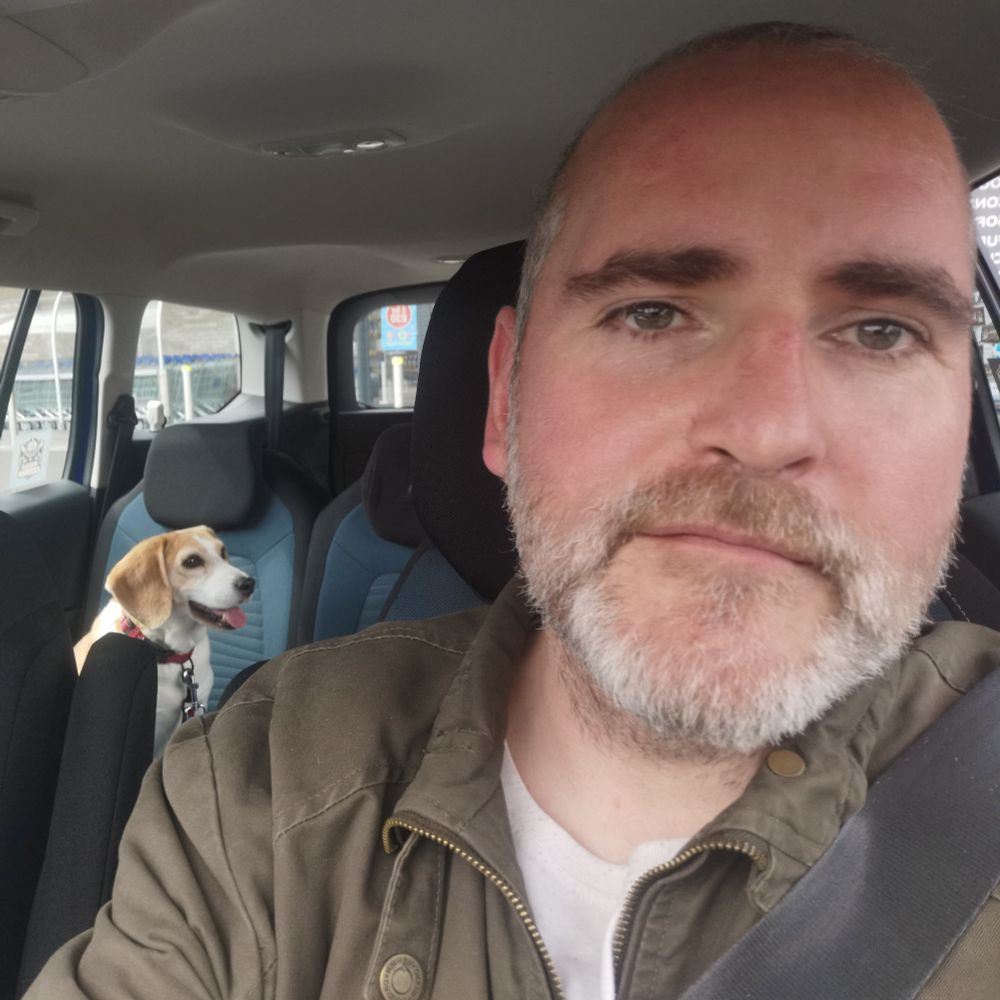 Me, with grey stubble, sitting in the driver's seat of the car. With Rio the tri beagle harnessed into the back seat. 