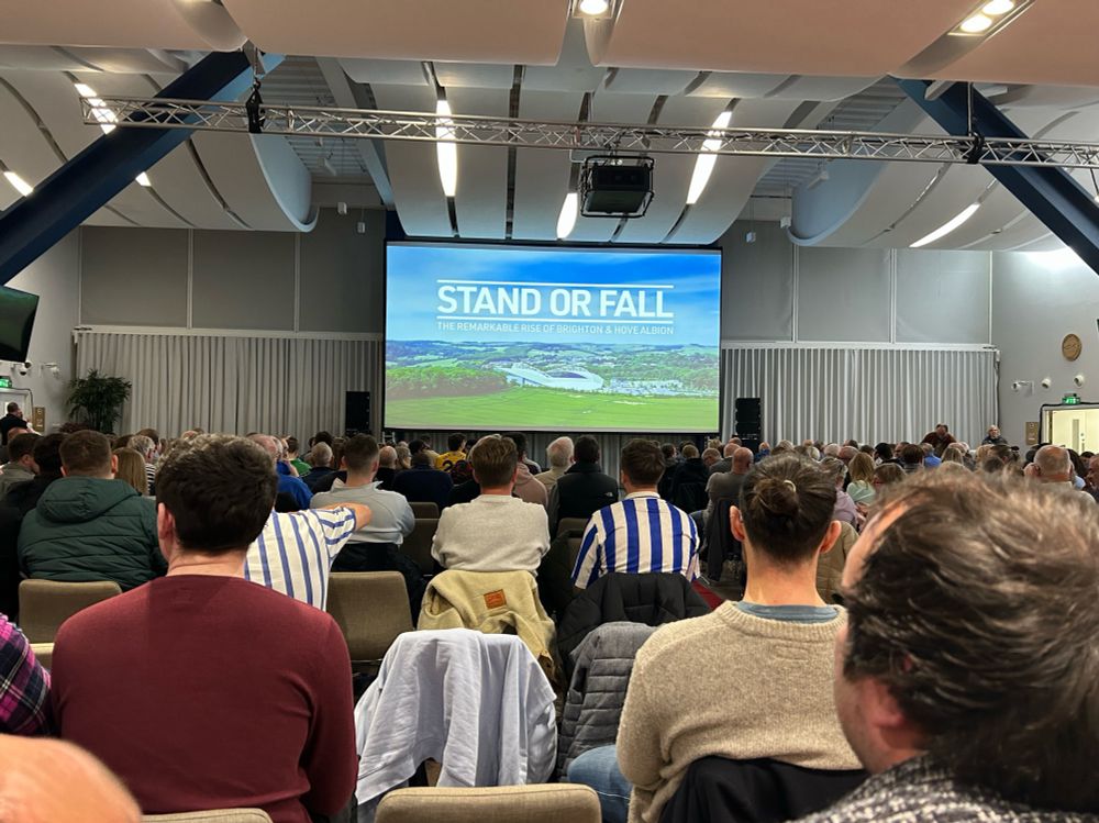 A large projector screen with a photo of the Amex Stadium and the words ‘Stand or Fall’  on top, in a large conference room with people sat on seats. 