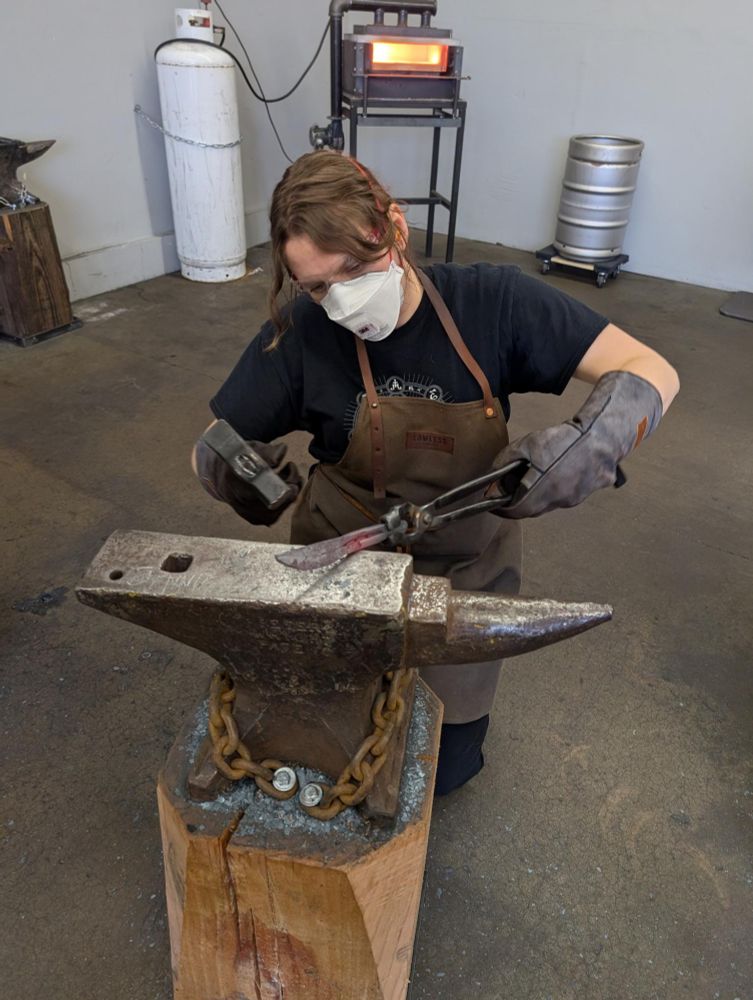A person knelt by an anvil with a forge in the background. They have a hammer and tongs holding a knife-shaped piece of metal in their hands.