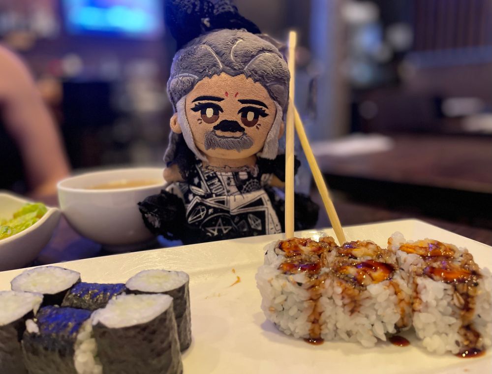 An Ed plushie by Rights is wearing a black and white tarot card dress with black ruffles and a matching hair bow. He has chop sticks that are bigger than him to the right. In front of him is a plate with cucumber rolls and sweet potato rolls