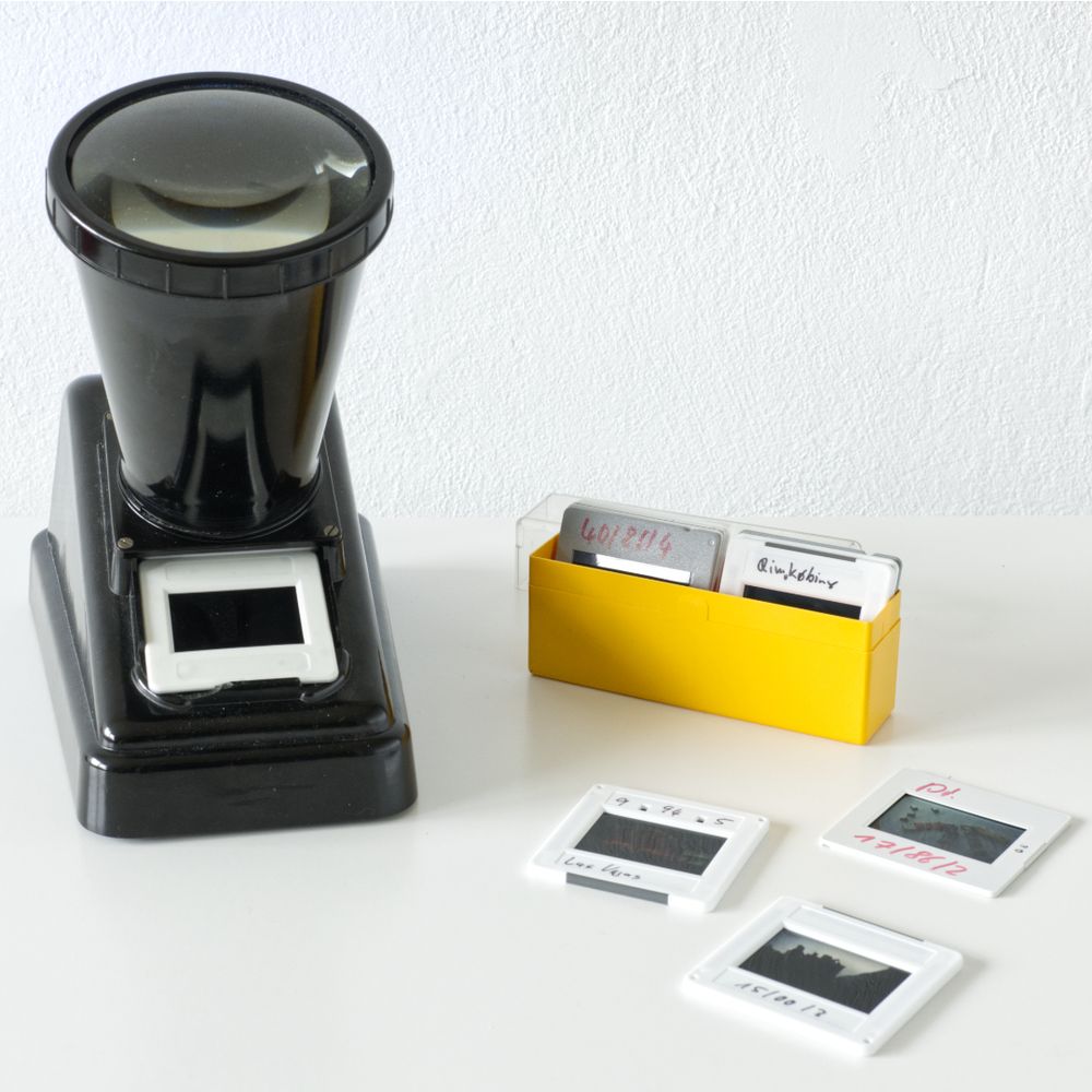 Quadratisches Farbfoto zeigt auf einem weißen Untergrund vor weißem Hintergrund einen altmodischen Diabetrachter mit eingelegten Dias. Daneben steht ein kleines gelbes Kästchen mit Dias und drei Dias liegen herum.
