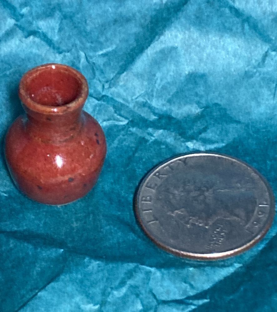 very small pottery vase. It's about as tall as a quarter, maybe two thirds the diameter. It's got a rounded base and a narrowed neck, and is suitable for putting small flowers in, that kind of thing. The barbies will find other uses, I'm sure. It has a dark red glaze with irregular black spots, and it's much prettier than my picture suggests. It's on the blue paper she wrapped it in, and next to a US quarter for scale. 
