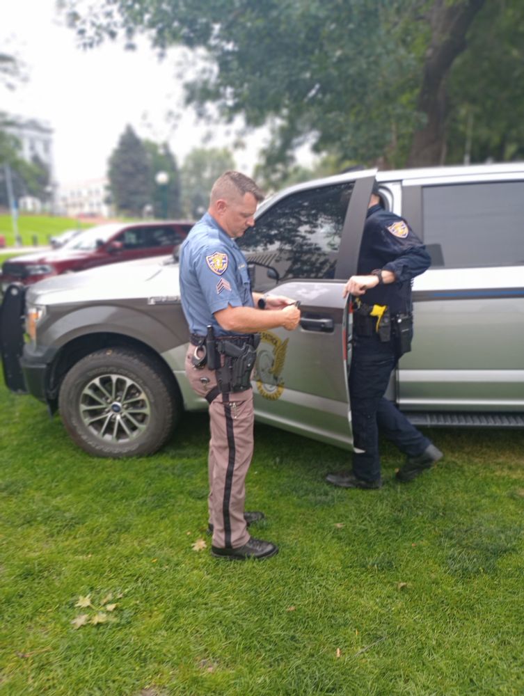 Two Colorado state troopers stand on the grass of Lincoln Veterans Memorial Park. A police SUV is parked behind them on the grass 