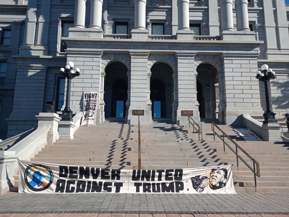 A banner reading DENVER UNITED AGAINST TRUMP in front of the Capitol steps 