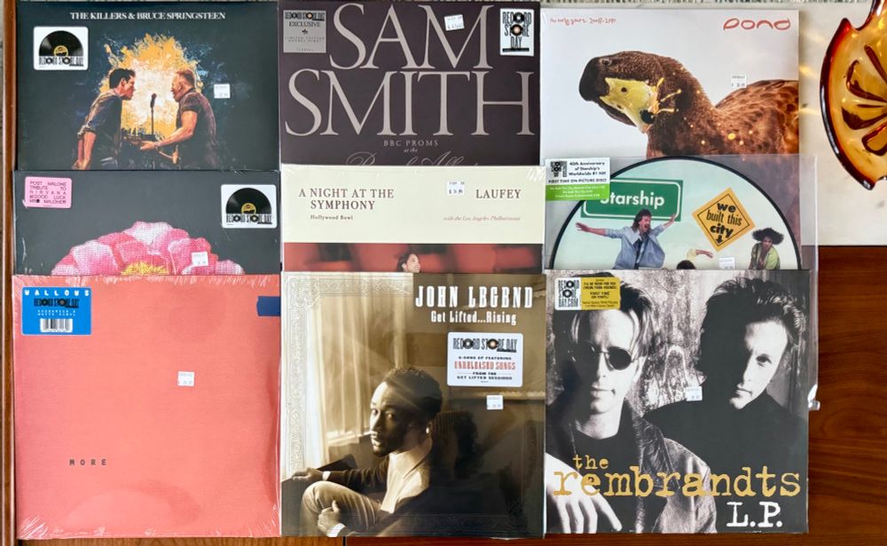Nine vinyl records from Record Store Day 2025 are displayed on a wooden table in three rows. Top row: The Killers & Bruce Springsteen single, Sam Smith ‘BBC Proms at the Royal Albert Hall,’ and Pond ‘The Early Years: 2008-2010.’ Middle row: Post Malone ‘Tribute to Nirvana,’ Laufey ‘A Night at the Symphony: Hollywood Bowl,’ and Starship ‘We Built This City’ 40th Anniversary picture disc. Bottom row: Wallows ‘More,’ John Legend ‘Get Lifted… Rising,’ and The Rembrandts ‘L.P.’ All records feature Record Store Day exclusive stickers.