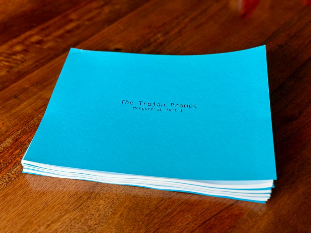 A bright blue manuscript titled The Trojan Prompt: Manuscript Parts 1-4 sits on a wooden table. The pages are neatly stacked and bound, with the title centered in black typewriter-style font on the cover page.