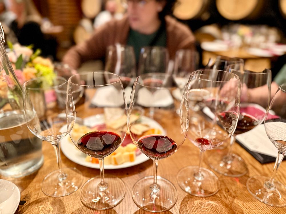 A wine tasting setup with several glasses of red wine arranged on a wooden table. In the background, blurred guests are seated, and there are cheese and charcuterie plates, floral centerpieces, and wine barrels lining the walls, suggesting a cozy winery setting.