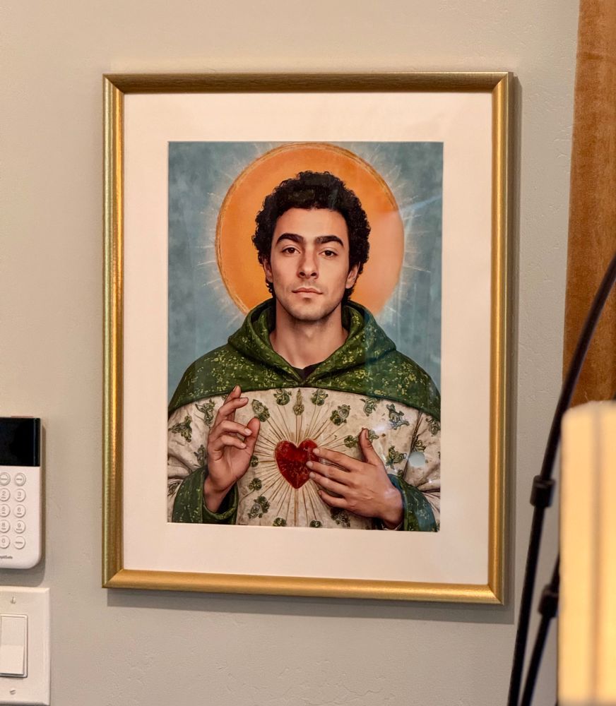 This is a framed portrait of Luigi Mangione depicted in a style reminiscent of traditional religious art, resembling Jesus Christ. He is shown with a radiant golden halo, wearing a green and white robe adorned with intricate floral patterns and a Sacred Heart symbol on his chest, emphasizing themes of devotion and spirituality, while the golden frame adds a classical and reverent touch to the piece.