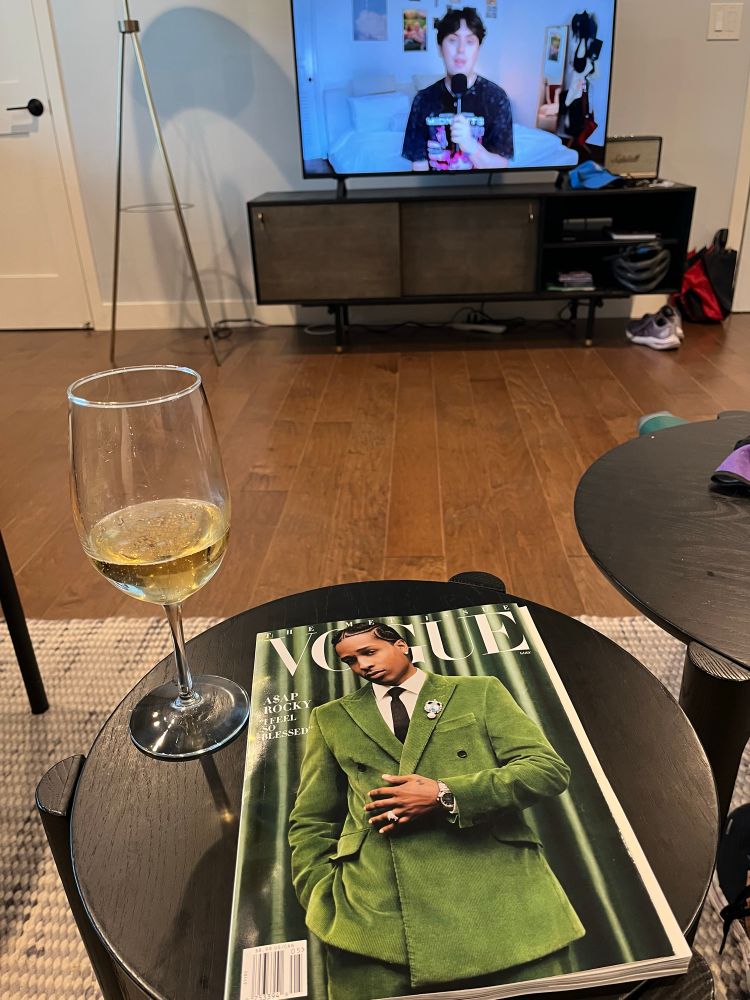 Looking out into a living room with a TV in the background and the Swiftologist YouTube channel playing.  In the foreground, a round coffee table with a glass of wine to the left and a Vogue magazine in the center.  The magazine cover features a handsome man in a kelly green corduroy suit.