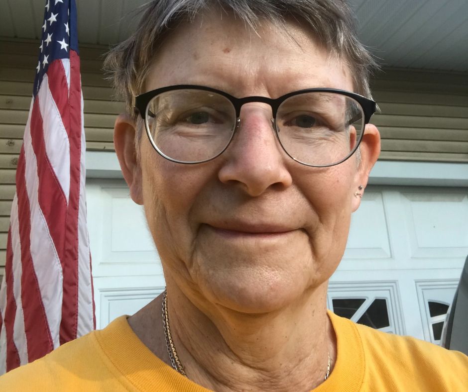 Susan is looking into the camera. She has on a yellow shirt. There is an American flag behind her.