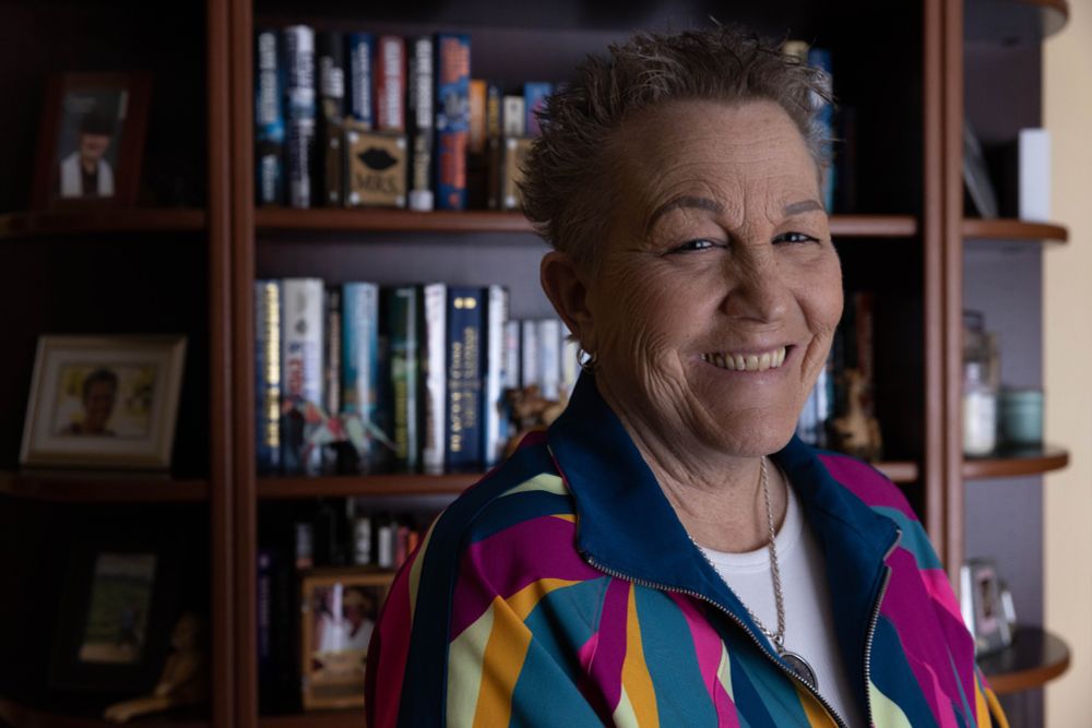 Photo of Deb in bright track suit. She is smiling at the camera and standing in front of a bookcase.
