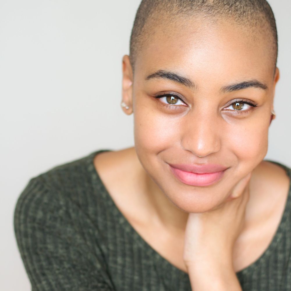 A portrait photo of Naomi, wearing a green shirt and smiling at the camera while holding her head in one hand.