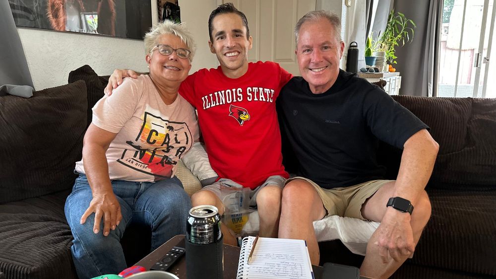 Andrew Flack sits on a couch between two people. They are all smiling at the camera. Andrew is wearing an Illinois State shirt.