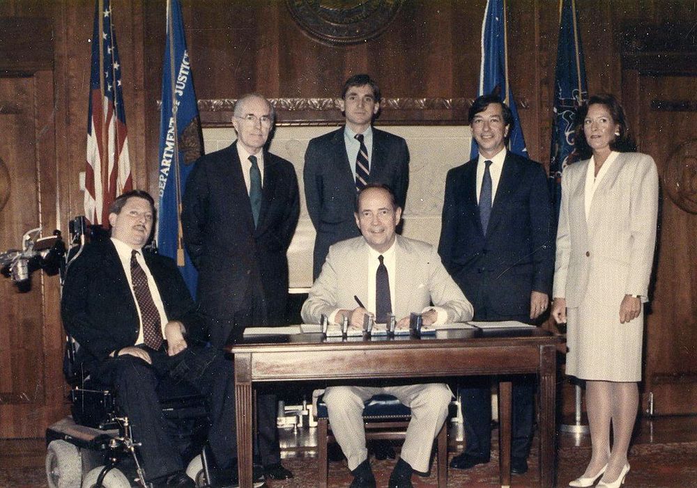 Andrew Batavia is at the Department of Justice. He is wearing a suit and tie, and sits in a motorized wheelchair to the left of a large desk. Behind the desk sits U.S. Attorney General Dick Thornburgh. Three men stand behind him and one woman stands to the right of the desk.