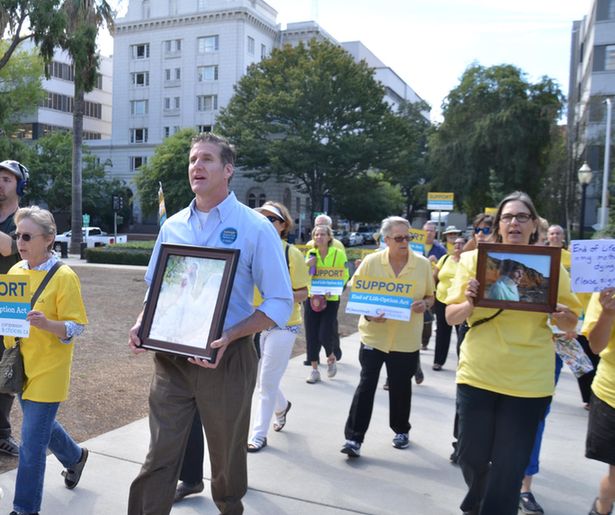 Advocates in yellow shirts march outside with Dan Diaz to support end-of-life options in California. Dan and several advocates hold framed photos of loved ones.