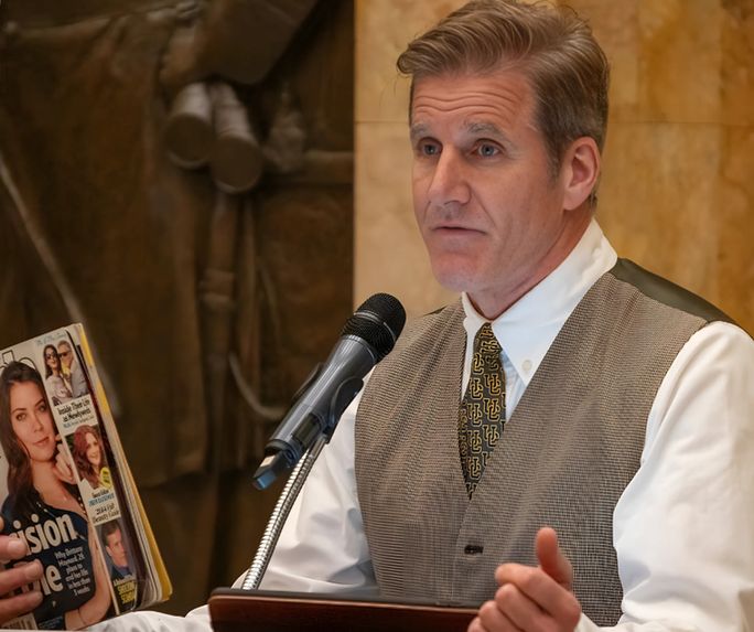 Photo of Dan Diaz speaking in front of a microphone and holding up a People magazine that has his late wife, Brittany Maynard, on the cover.