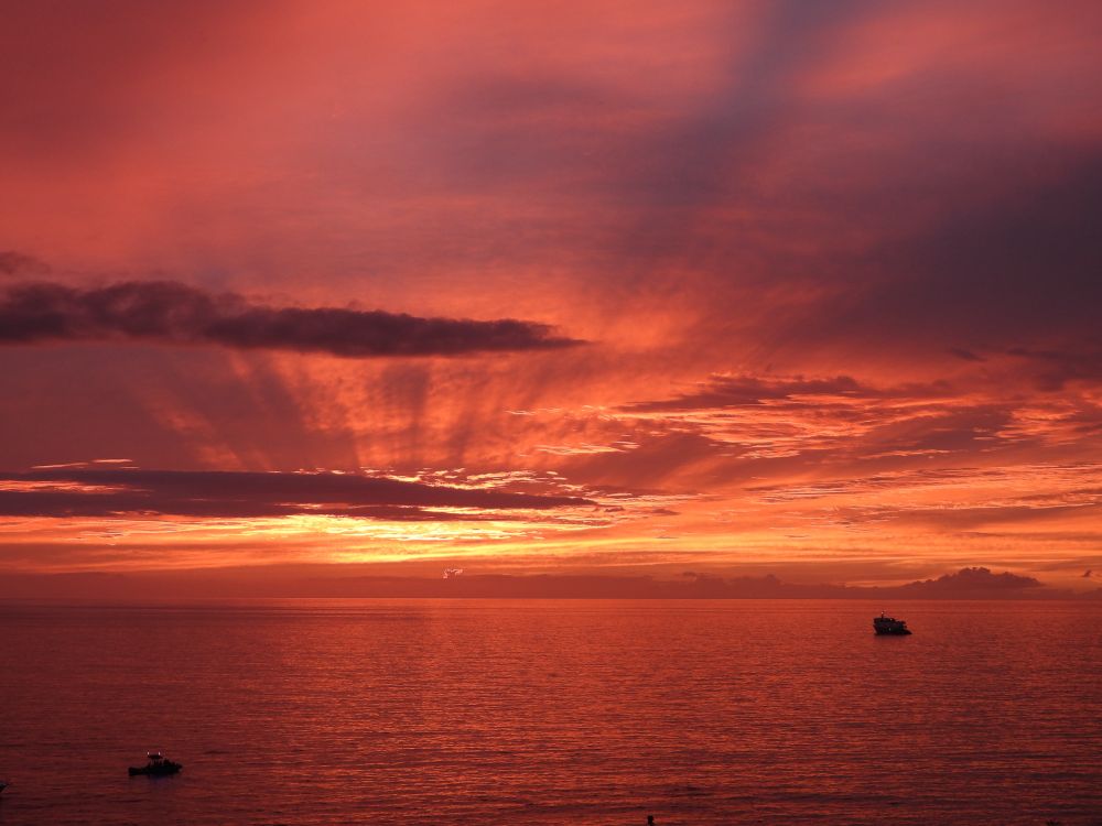 An amazing orange and gold sunset fills the sky over the Pacific. Two boats are silhouetted in the foreground. 