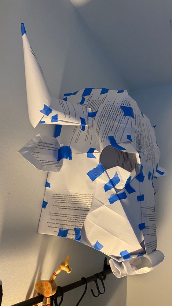 A sculpture of the head of a bison made with folded and cut scrap paper, and bits of blue painter’s tape, hanging on a wall.
