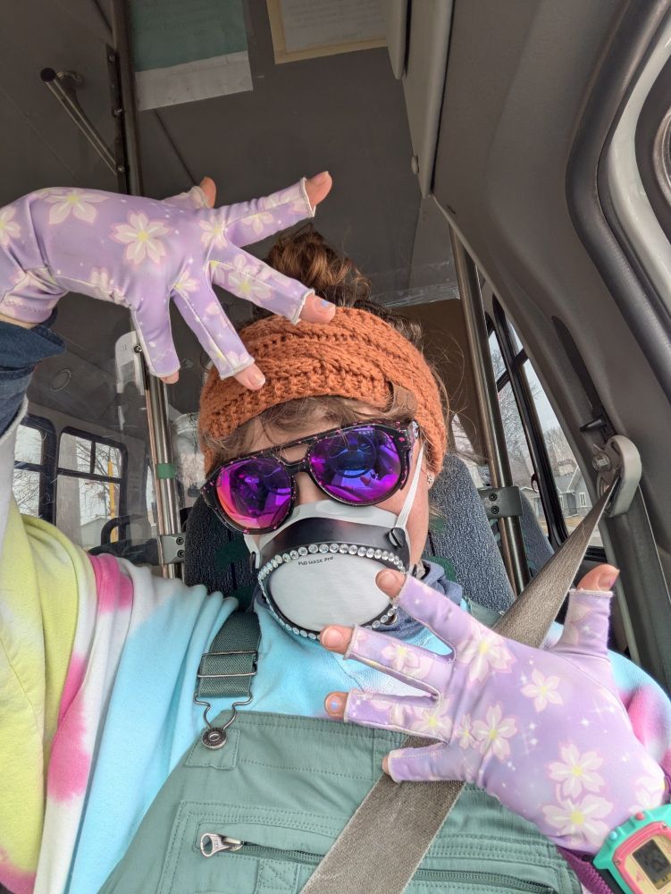 Similar to the last photo, a selfie of Syd, a white person wearing purple sunglasses, a burnt orange headband, a FloMask with sparkly stickers around the edge, a tie dye shirt, and green overalls. Syd holds up their hands in a face framing jazz hands kinda way, showing off the purple flowery compression gloves they have on. 