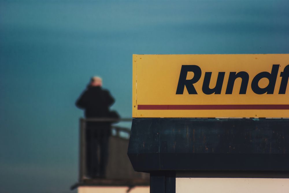 Foto eines unscharf in der Ferne auf einem Balkon stehenden Mannes, im Vordergrund ein Schild, abgeschnitten der Schriftzug "Rundflüge"