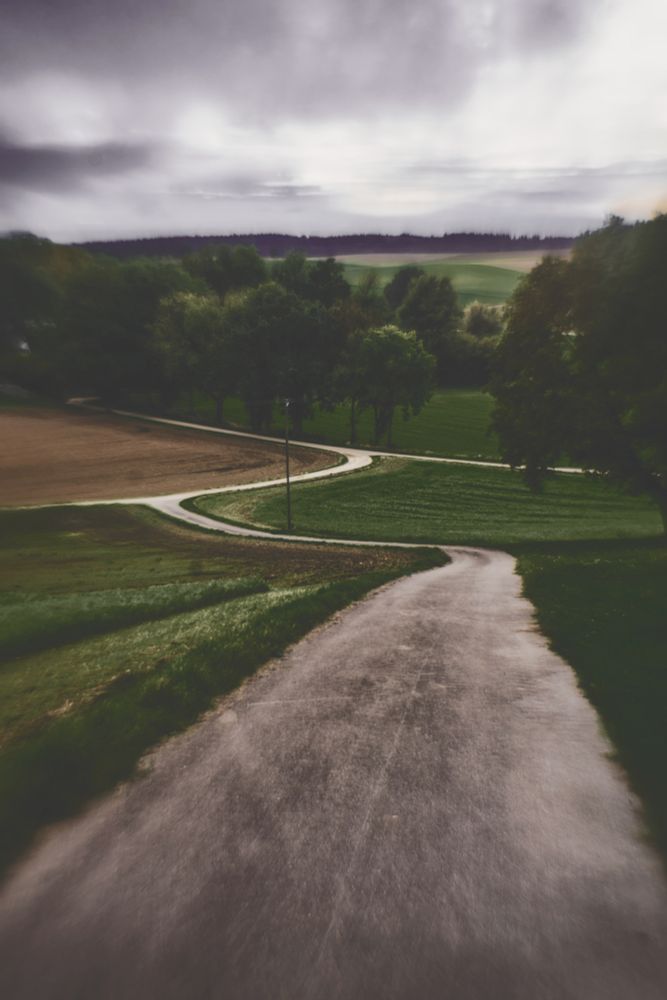 An den Rändern unscharfes Foto einer Landschaft durch die sich ein Teerweg schlängelt. Felder, Bäume, ein Strommast