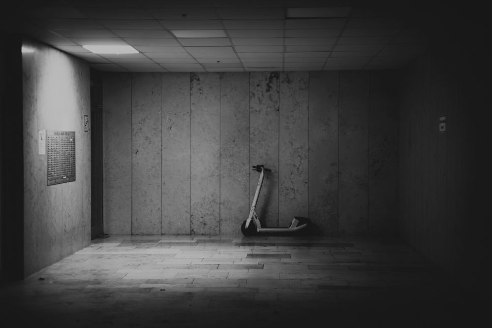 Schwarzweissfoto: Ein Hauseingang eines großeren Hauses, spärlich beleuchtet, hinten steht ein E-Roller
