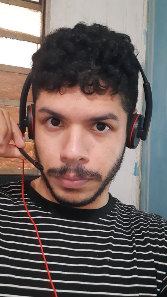 Homem pardo (ou preto de pele clara) com cabelo e bigode de cor preta, com camisa listrada preta e branca e fones de ouvido com microfone