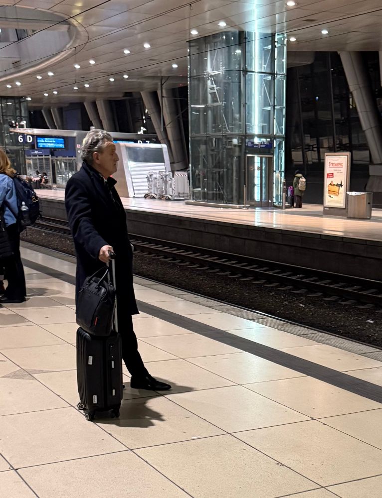 Frank N. an einem Bahngleis wartend mit einem Koffer.
