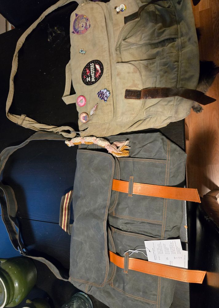 Two messenger bags laying side by side on a black table. The bag on the right is very worn and has a few pins and patches on it. The bag on the left is brand new, clean and holds a better shape