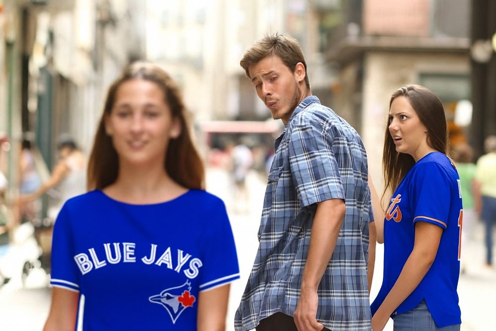 A “Distracted Boyfriend” meme scene on a sunny pedestrian street. In the foreground, a smiling woman in a bright blue Toronto Blue Jays jersey walks past, slightly out of focus. In the middle, a man in a short-sleeved blue plaid shirt turns his head to look at her with raised eyebrows. On the right, a woman in a royal blue New York Mets jersey and matching Mets baseball cap looks at him with wide-eyed, incredulous surprise. The background is softly blurred with buildings and pedestrians.