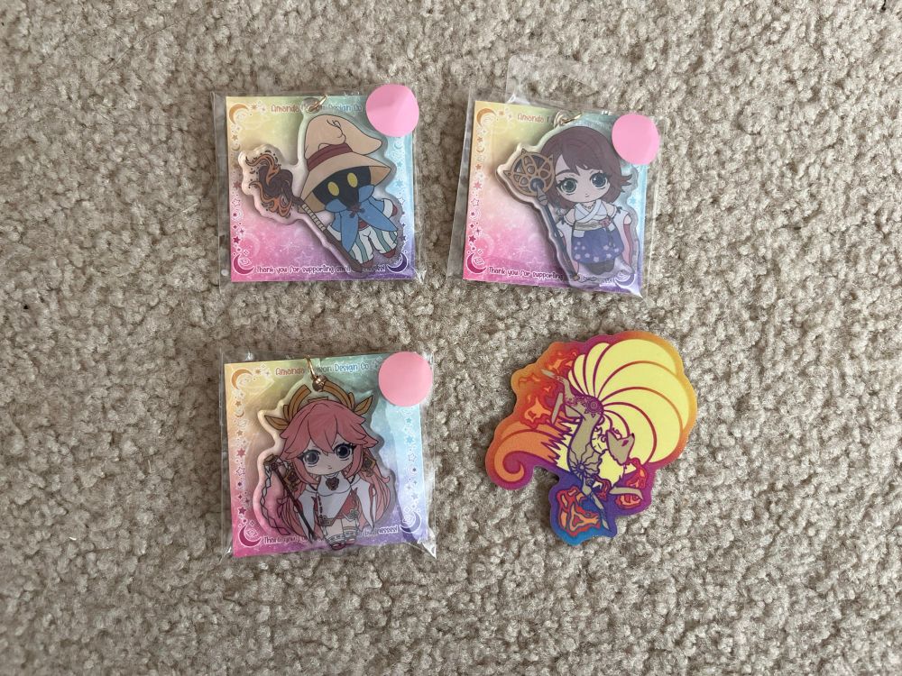Merch from Amanda Nelson Design, consisting of: keychains of Vivi from FFIX, Yuna from FFX, and Yae Miko from Genshin Impact; a colorful sticker of Ninetales the Pokémon .