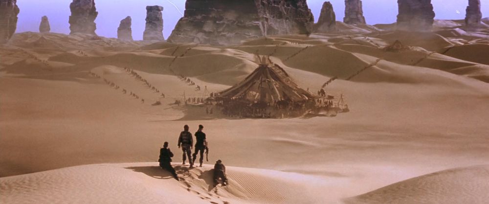 A group of four characters standing on top of a sand dune, looking at a city in the distance.